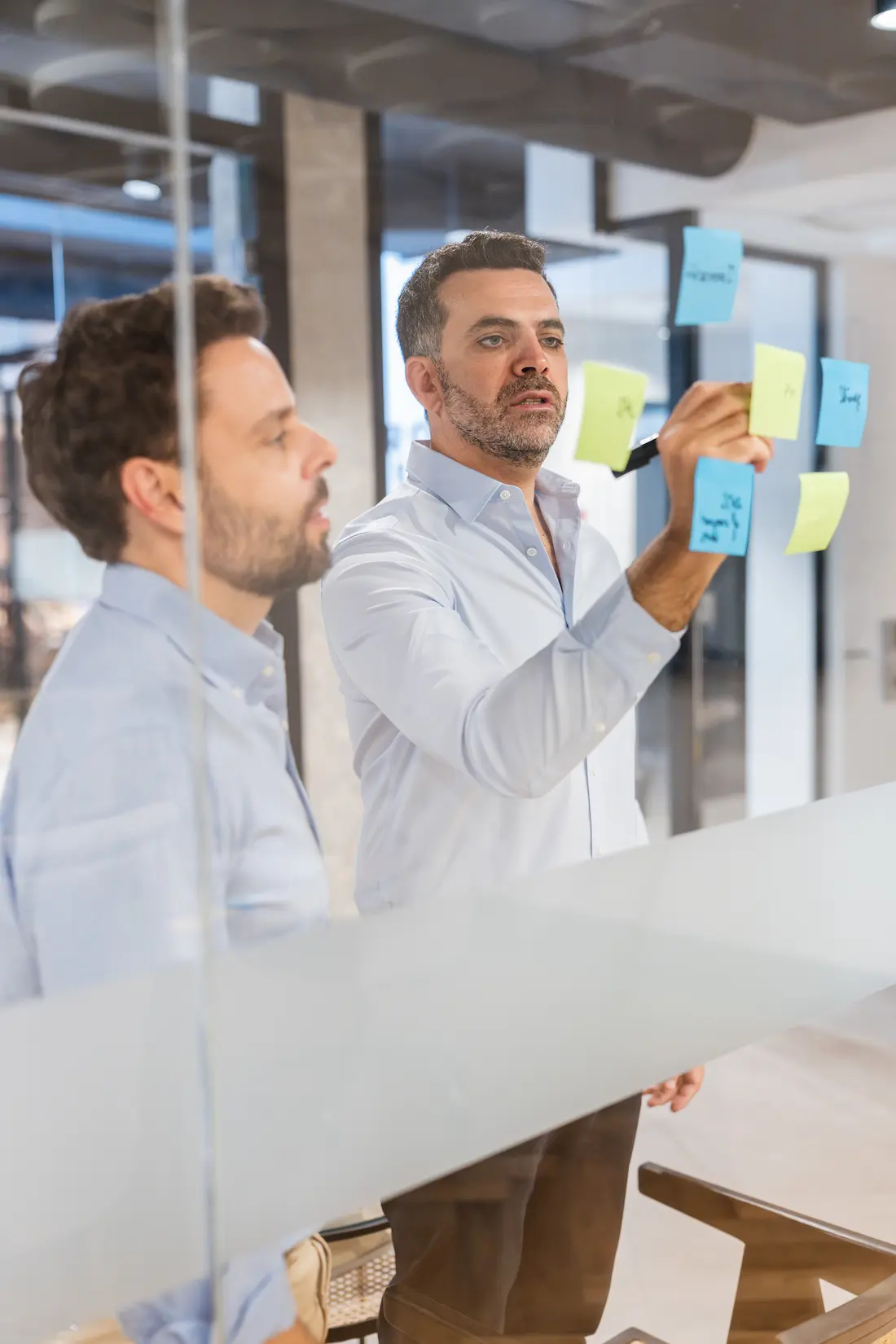 Two colleagues placing colourful sticky notes on a glass wall during a strategy session